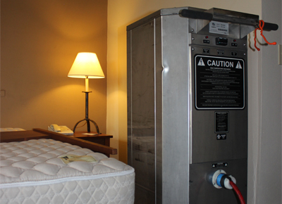 The image shows a professional heat treatment device positioned next to a hotel bed with a lamp, used for pest control to eliminate bed bugs through high-temperature treatment.